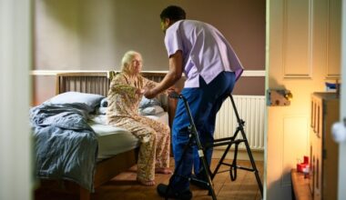 Senior woman in floral pajamas smiling and holding hands with a male carer, preparing to stand up from her bed.
