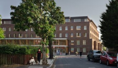 Appleby Blue Almshouse, a modern brick residential building with large windows, seen from across the street with pedestrians and trees in front.