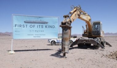 A sign reading &quot;First of its kind. HyIron Oshivela – Green Iron from Namibia&quot; stands next to an excavator breaking ground in a desert landscape.