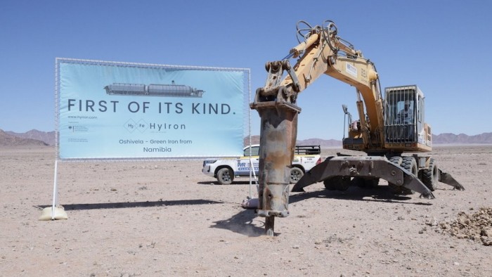 A sign reading &quot;First of its kind. HyIron Oshivela – Green Iron from Namibia&quot; stands next to an excavator breaking ground in a desert landscape.