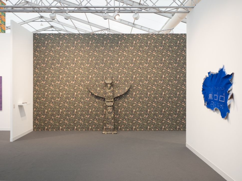 Booth featuring a wall covered in floral-patterned wallpaper with a totem-like sculptural figure at the center, and an abstract blue hide-shaped artwork on the side wall.