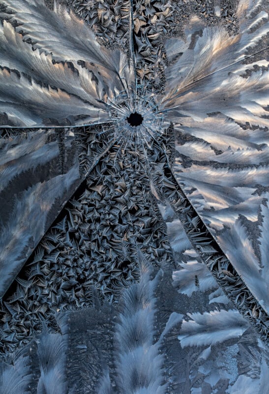 Close-up of intricate frost patterns radiating outward from a small central hole on a glass surface, with silvery and bluish hues creating a textured, almost feather-like appearance.