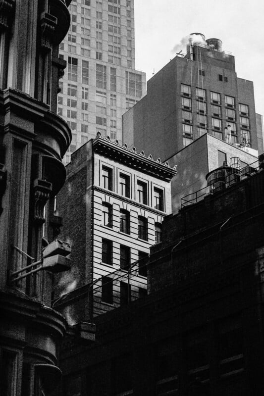 Black and white photo of tall buildings in a city, showing a mix of architectural styles and details, with shadows and smoke rising from a rooftop in the background.
