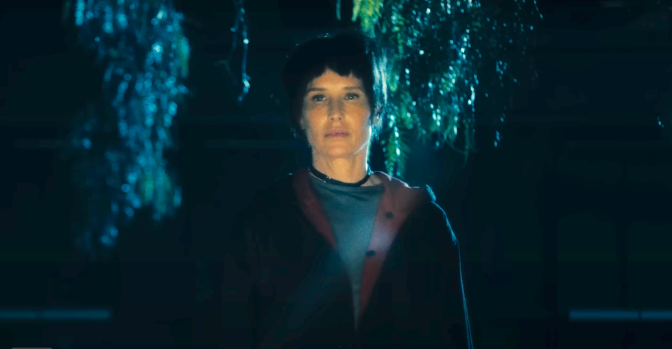 shawnee smith as amanda young in saw x, a woman with short black hair stands in a cave wearing a green top and red hoodie