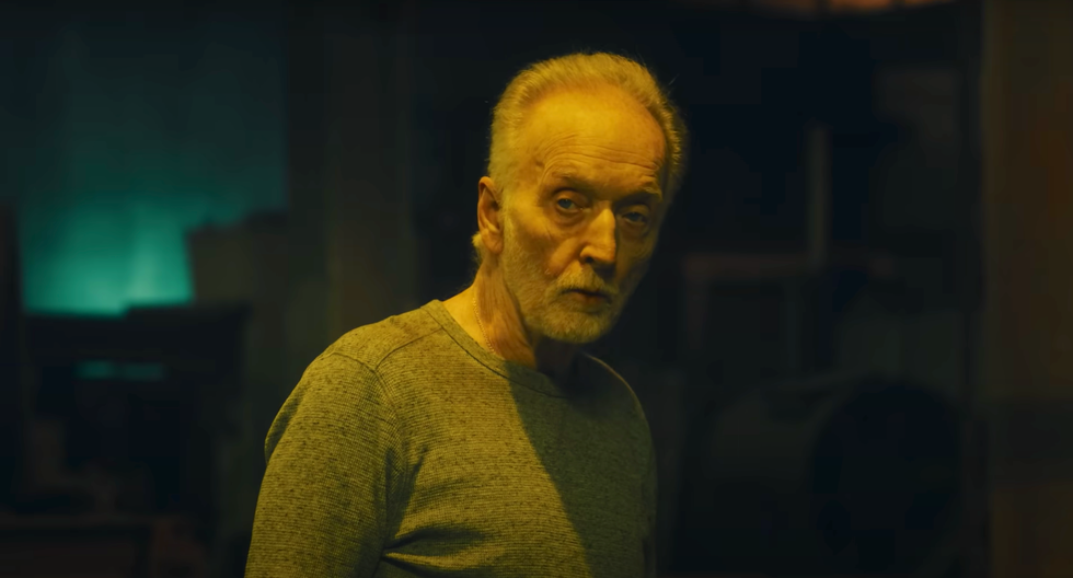 tobin bell as jigsaw in saw x, an older man with grey hair and beard stands in a dark room looking at the camera, he wears a long sleeve grey top