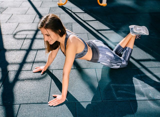 A woman is doing sports exercises. Knee push-ups. Workout for arms and pectoral muscles. Fitness for weight loss in open air sportsground. Light and shadow. Sunny morning.