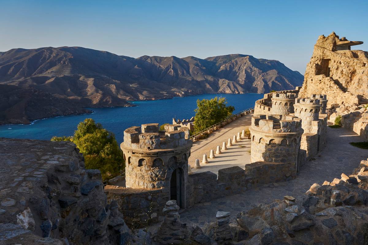 A view of blue water surrounding a historic piece of land in spain with mountains in the background