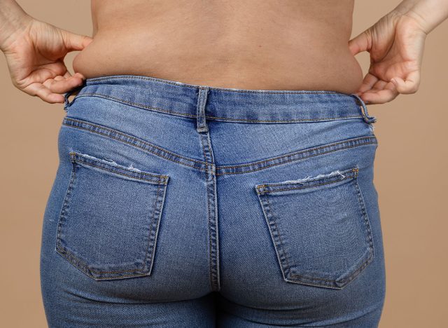 Closeup of young woman overweighted waist wearing blue jeans holding fat sides with hands. Body positive. Accepting who you are. Tight clothes. Need for wardrobe change.