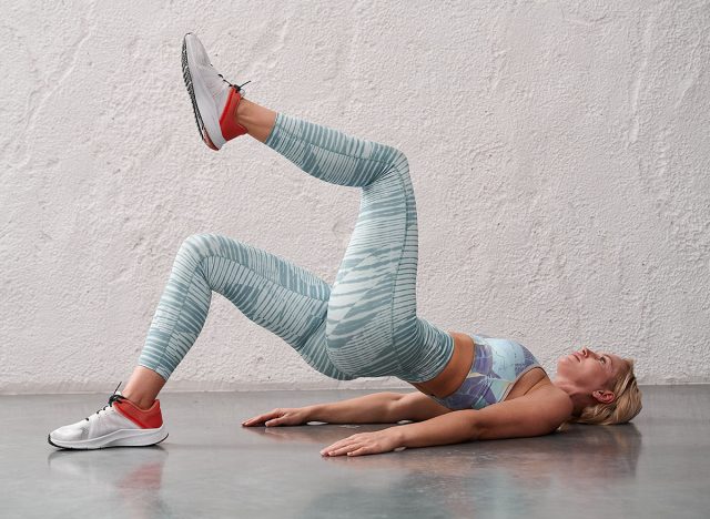 Young woman in sportswear performs a buttock bridge with one leg raised upand bent at the knee. Pilates studio. Young sportive woman doing a glute bridge isolated on gray background.