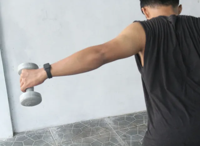 An Asian teenager is lifting small dumbbells to train his back muscles or bent over reverse fly in his backyard. #SSTKWellness