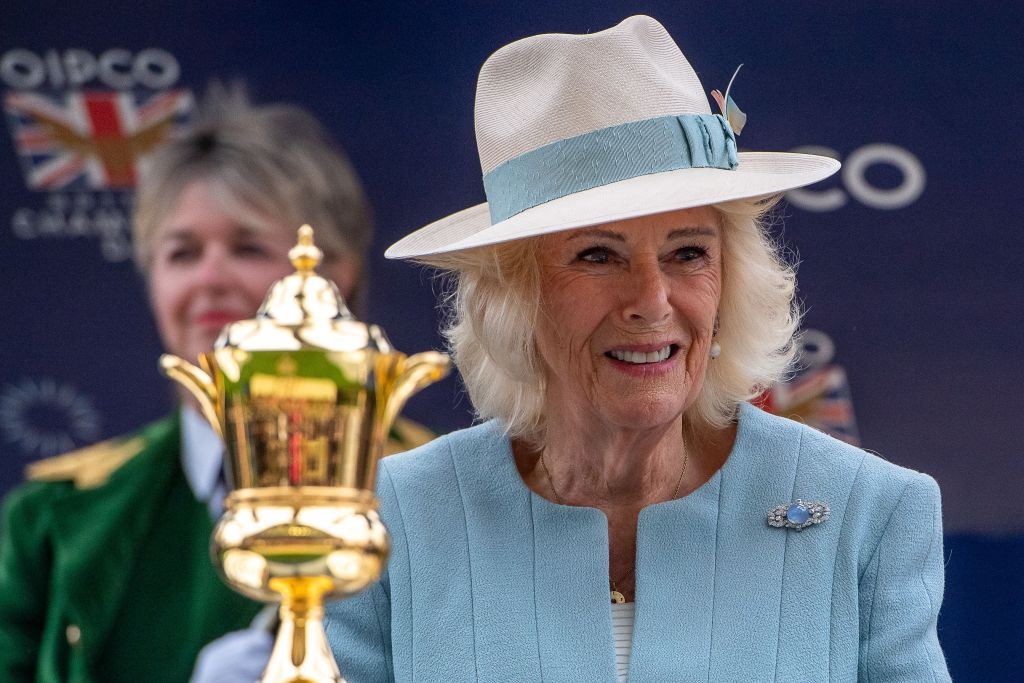 Queen Camilla smiling beside a gold trophy at Ascot, wearing a hat, blue jacket and a diamond brooch.