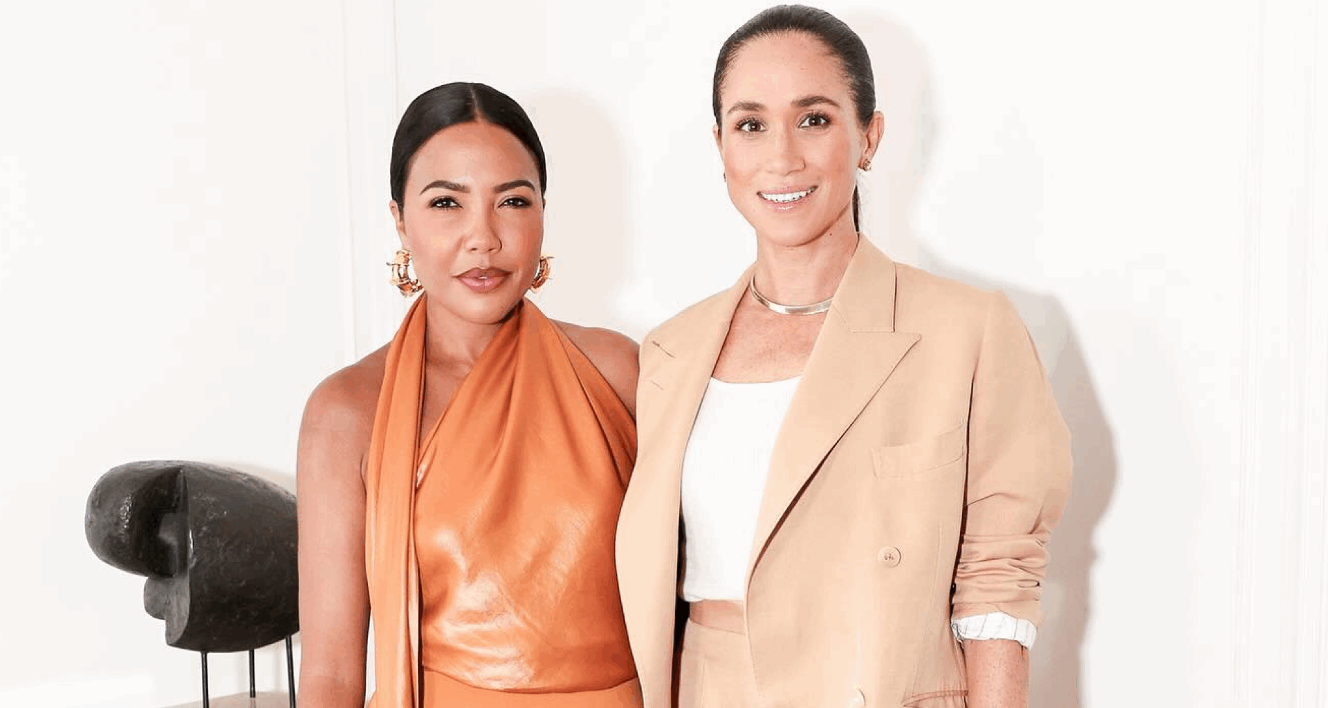 Meghan Markle wearing a beige suit standing with Emma Grede in an orange dress