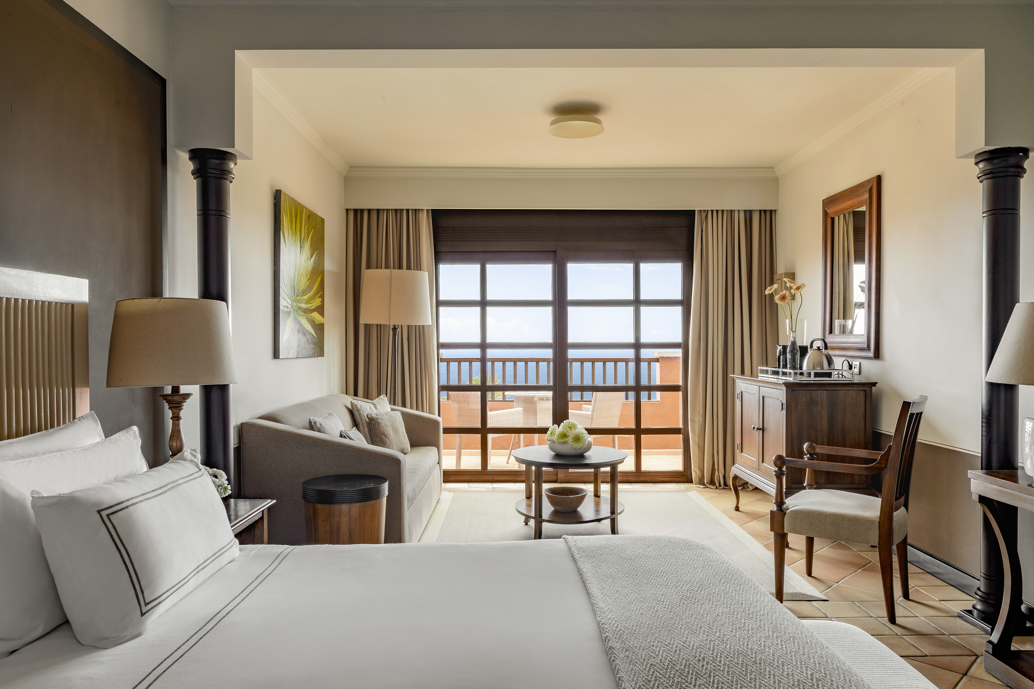 A hotel room with a bed, a sofa, a cabinet, and a large window looking out onto a patio and the ocean.