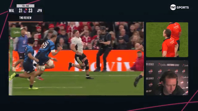 A rugby match showing a player in a white and black jersey running with the ball, while other players in blue jerseys follow. The score is Wales 21, Japan 23, with a "TMO REVIEW" message displayed.