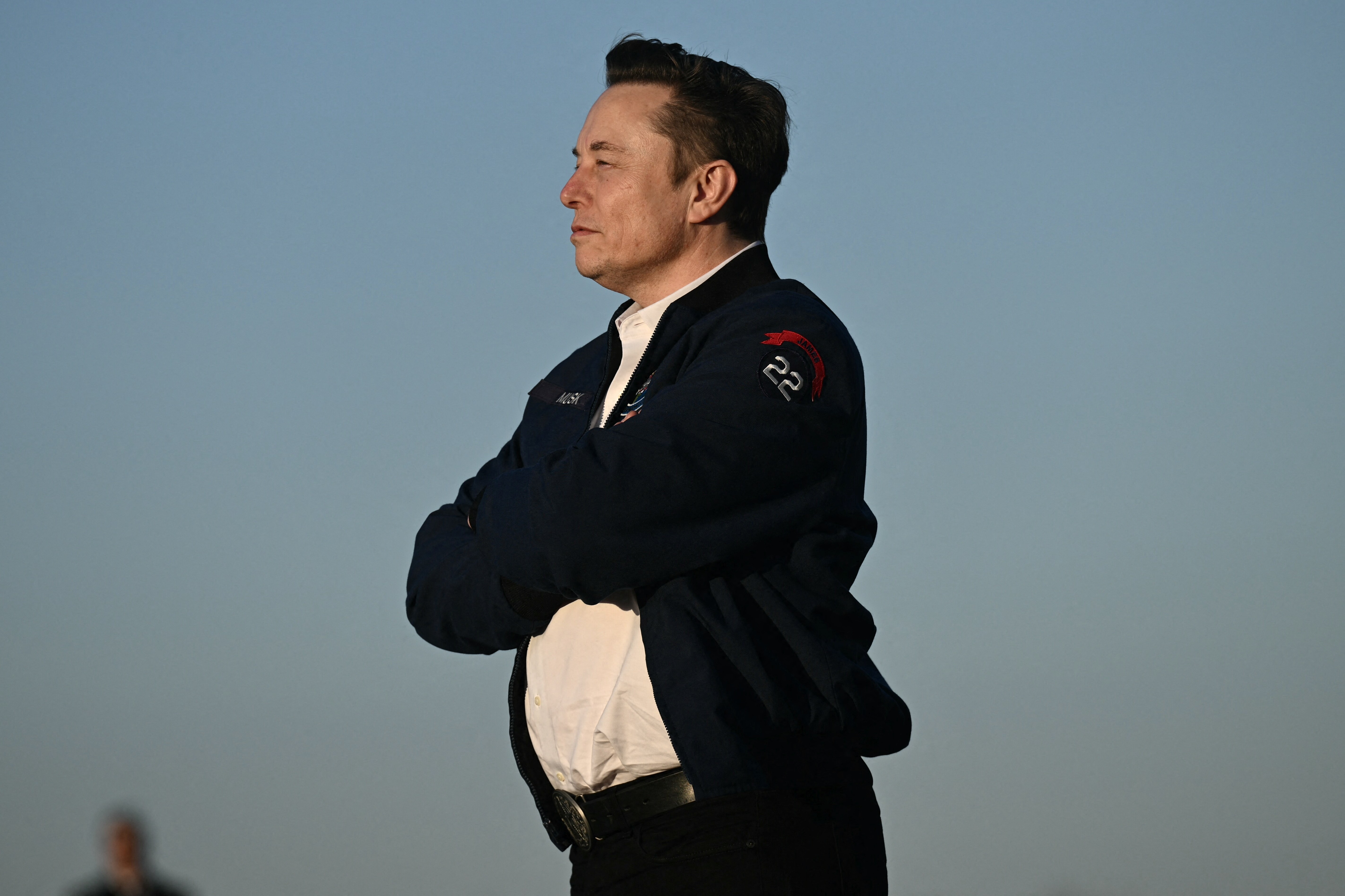 Elon Musk in a dark blue jacket and white shirt, looking to the right with arms crossed, against a clear blue sky.