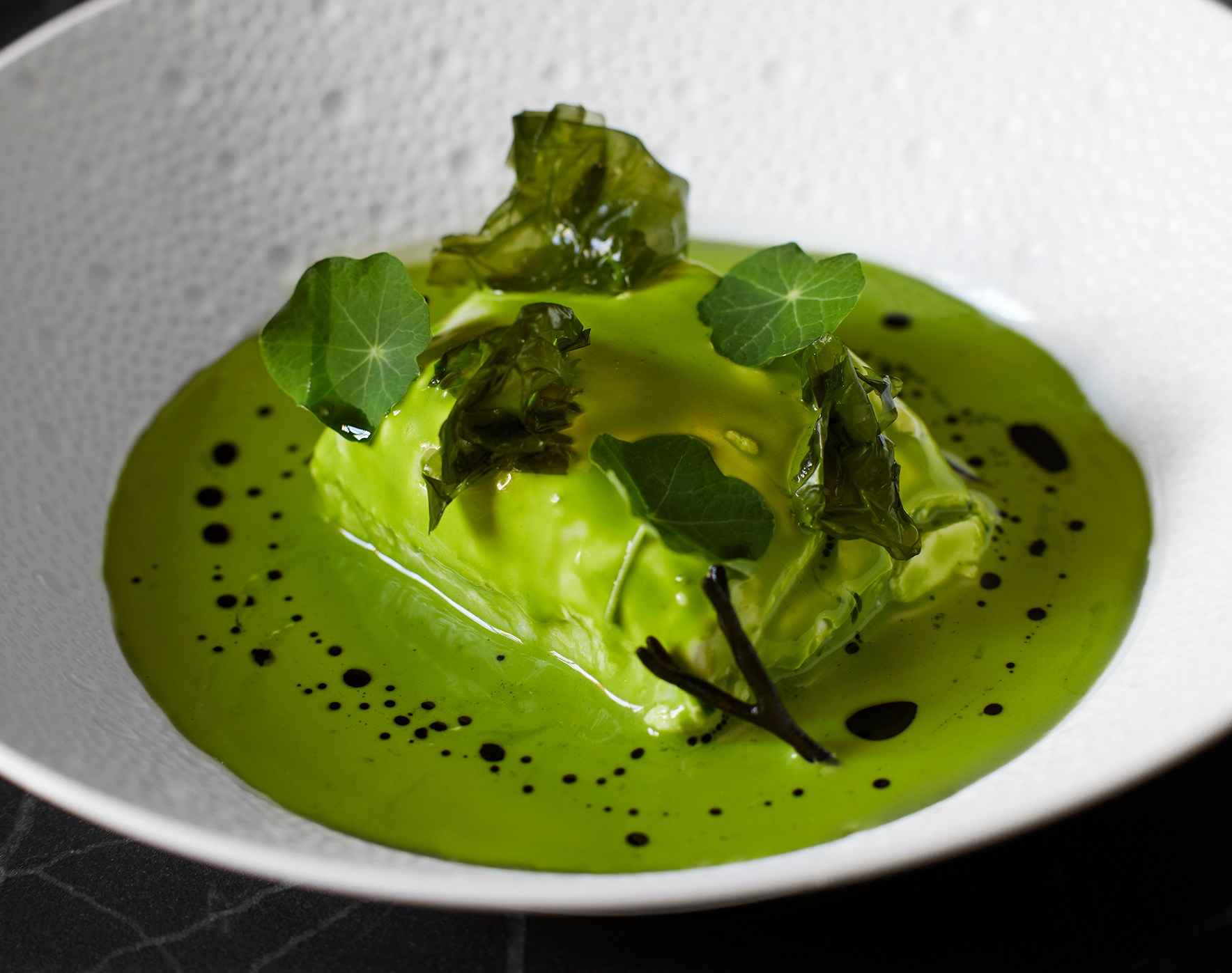 A bowl of bright green soup with dark green herbs and a garnish in the center.
