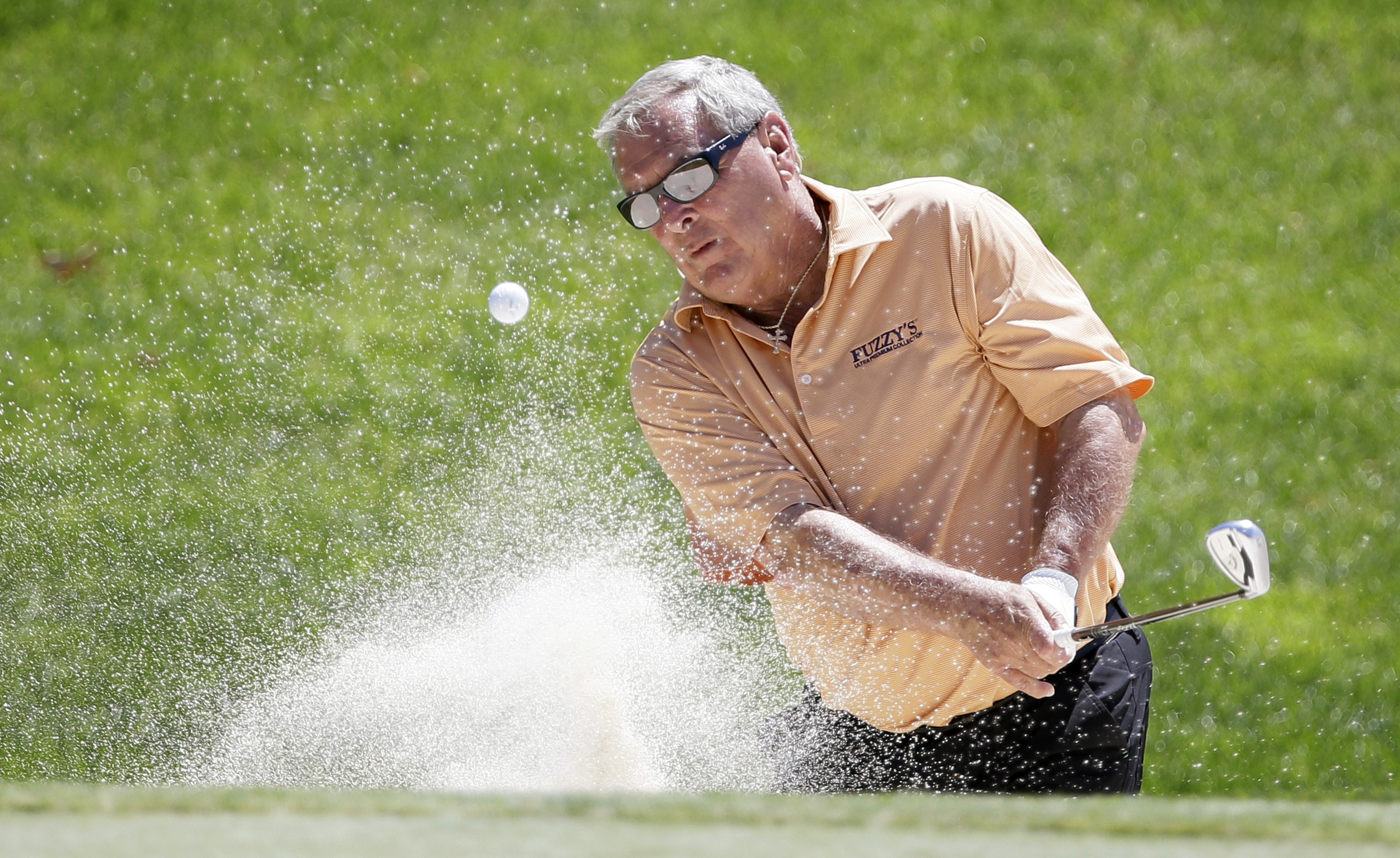 Fuzzy Zoeller hits a golf ball out of a bunker, scattering sand and dust into the air.