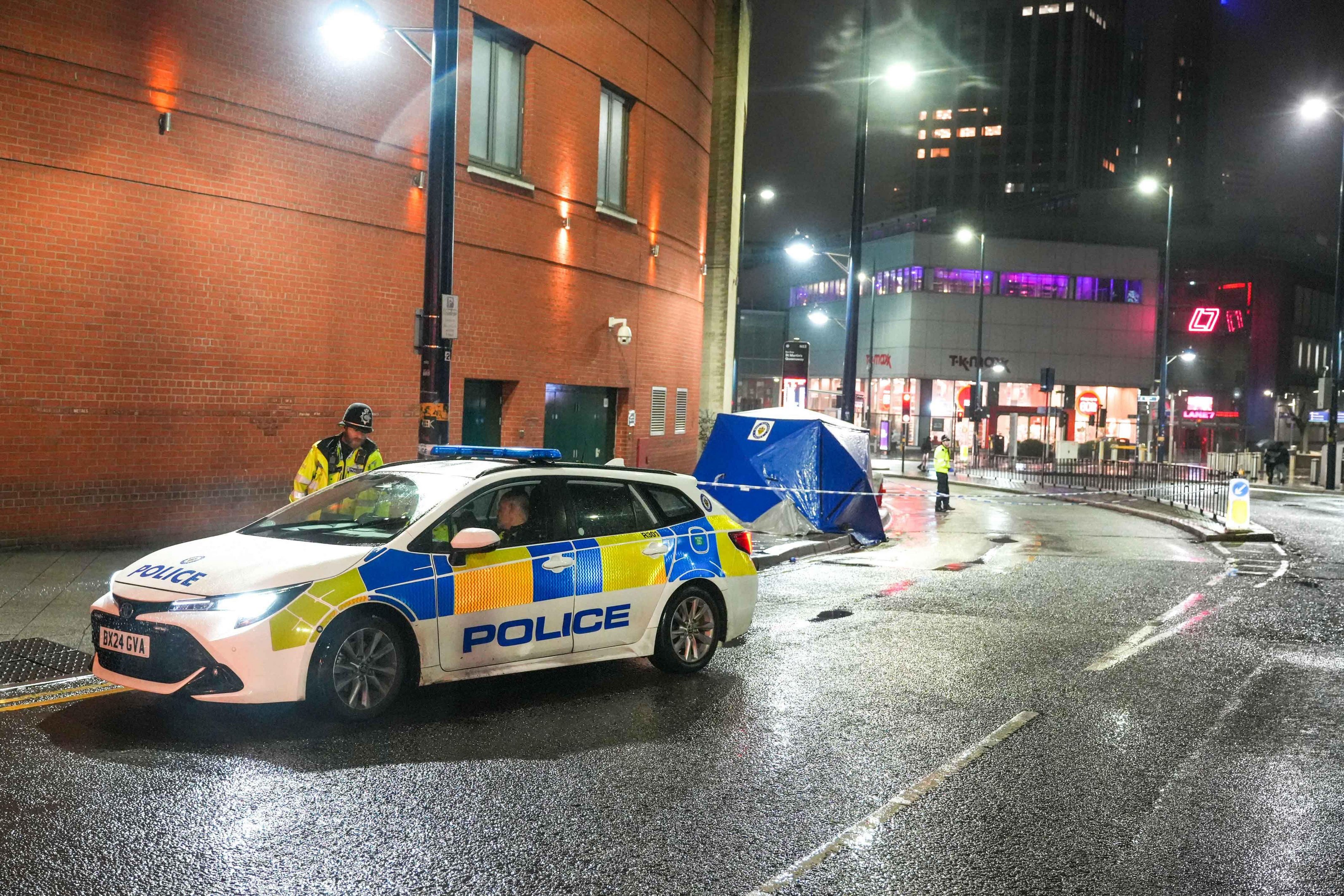 Police car and two officers at a crime scene with a forensic tent on a wet city street at night.