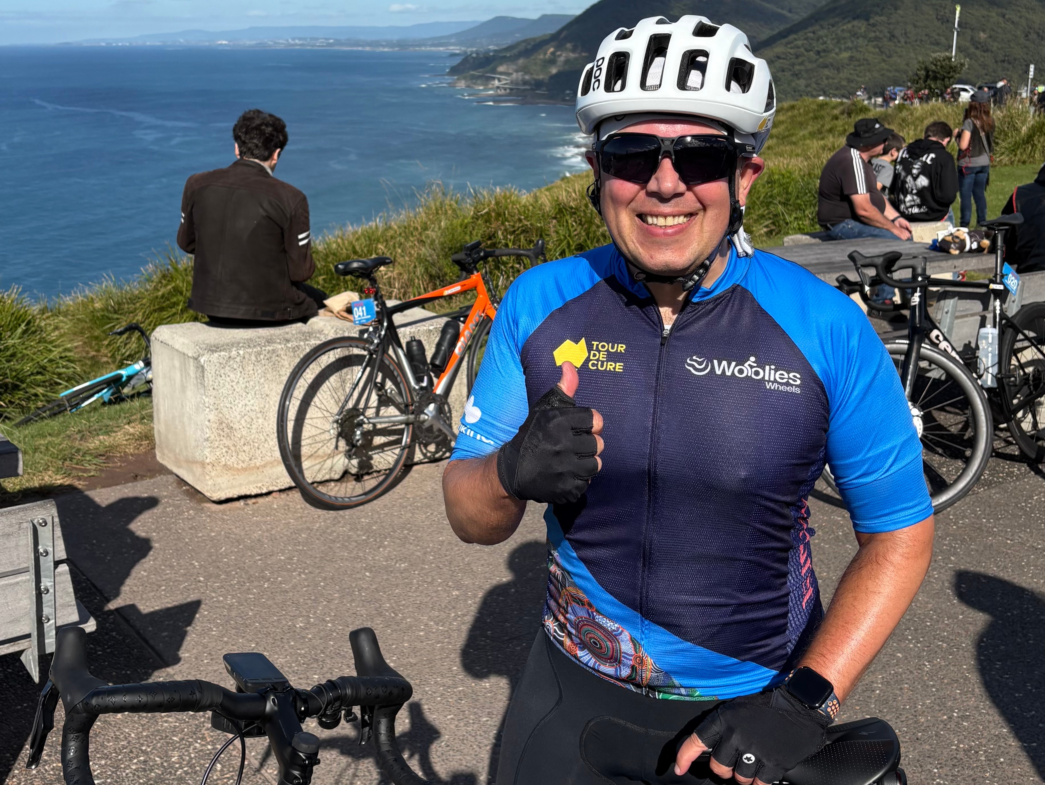 Cyclist Jon Clark in a helmet and sunglasses, wearing a "Tour De Cure" cycling jersey, gives a thumbs-up next to his bike with the ocean and hills in the background.