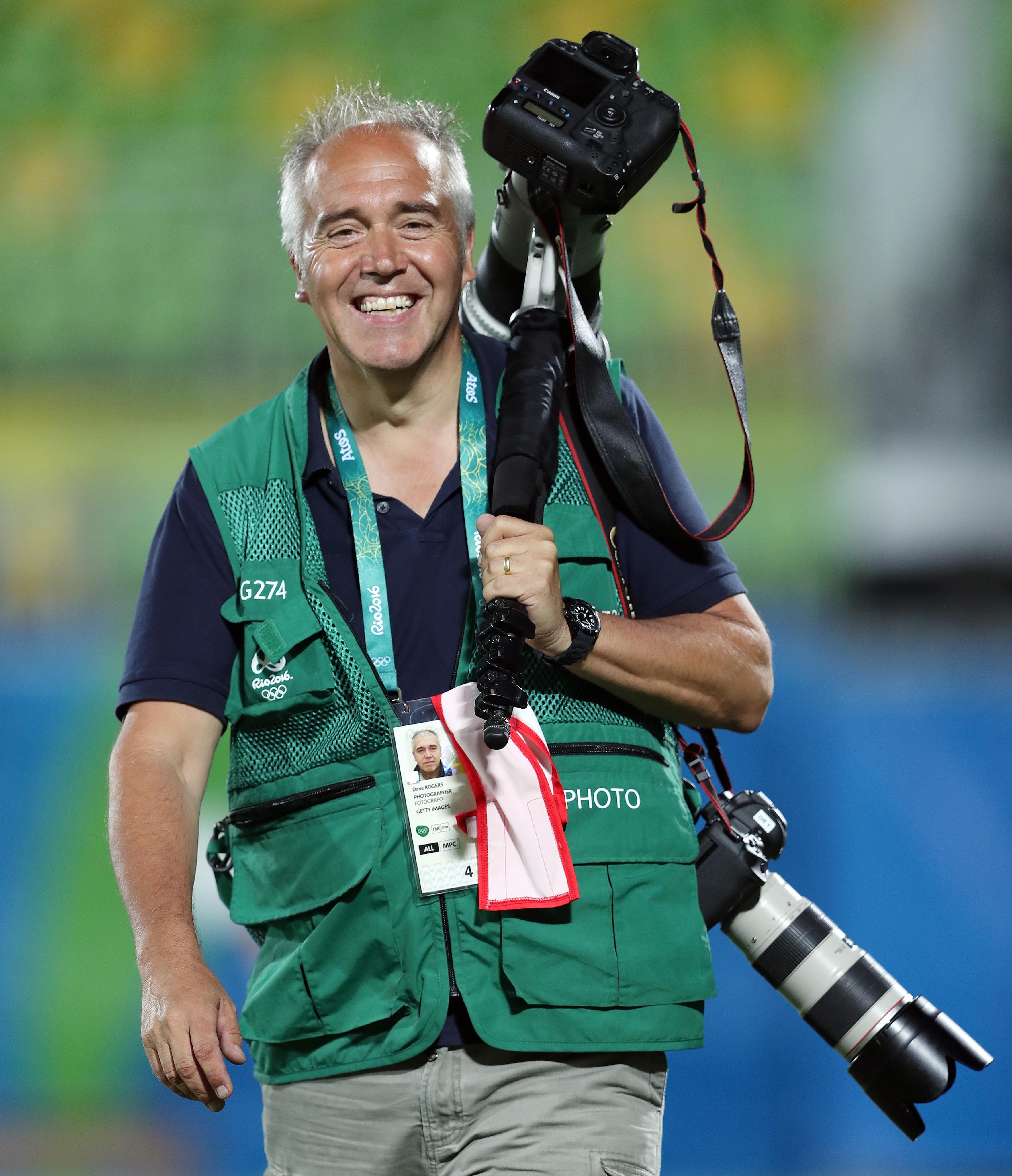 A male photographer wearing a green vest smiles while carrying a large camera with a long lens over his shoulder.