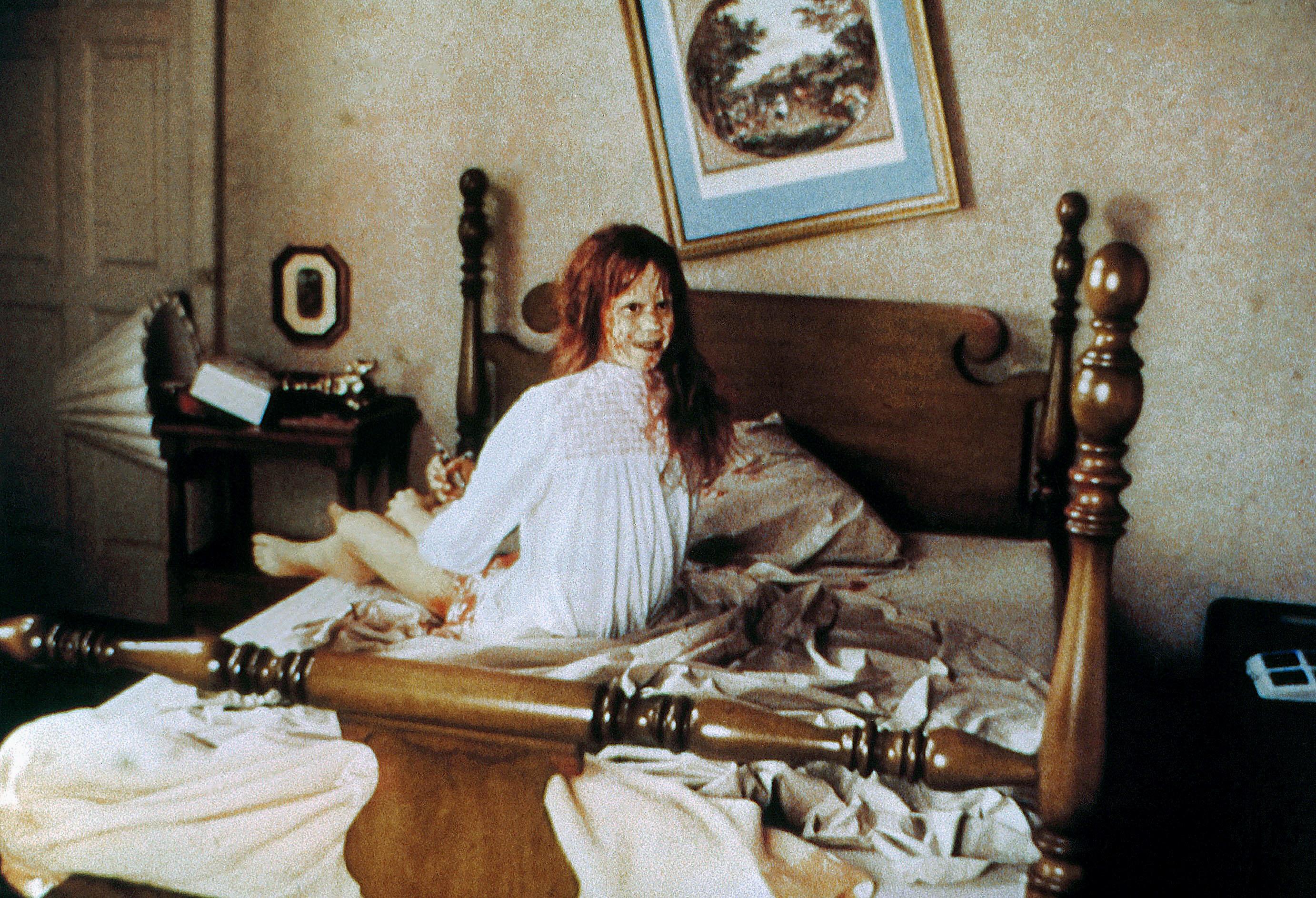 Linda Blair as Regan MacNeil from the film "The Exorcist," wearing a white nightgown covered in red, sitting up in a wooden bed.