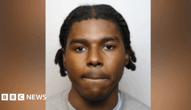 Young 18 year old man sits in the passenger seat of a black VW car, his arm resting on the open window as he looks to camera