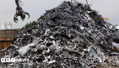 A huge pile of silver scrap metal, with a metal crane claw coming down towards it, to grab it.