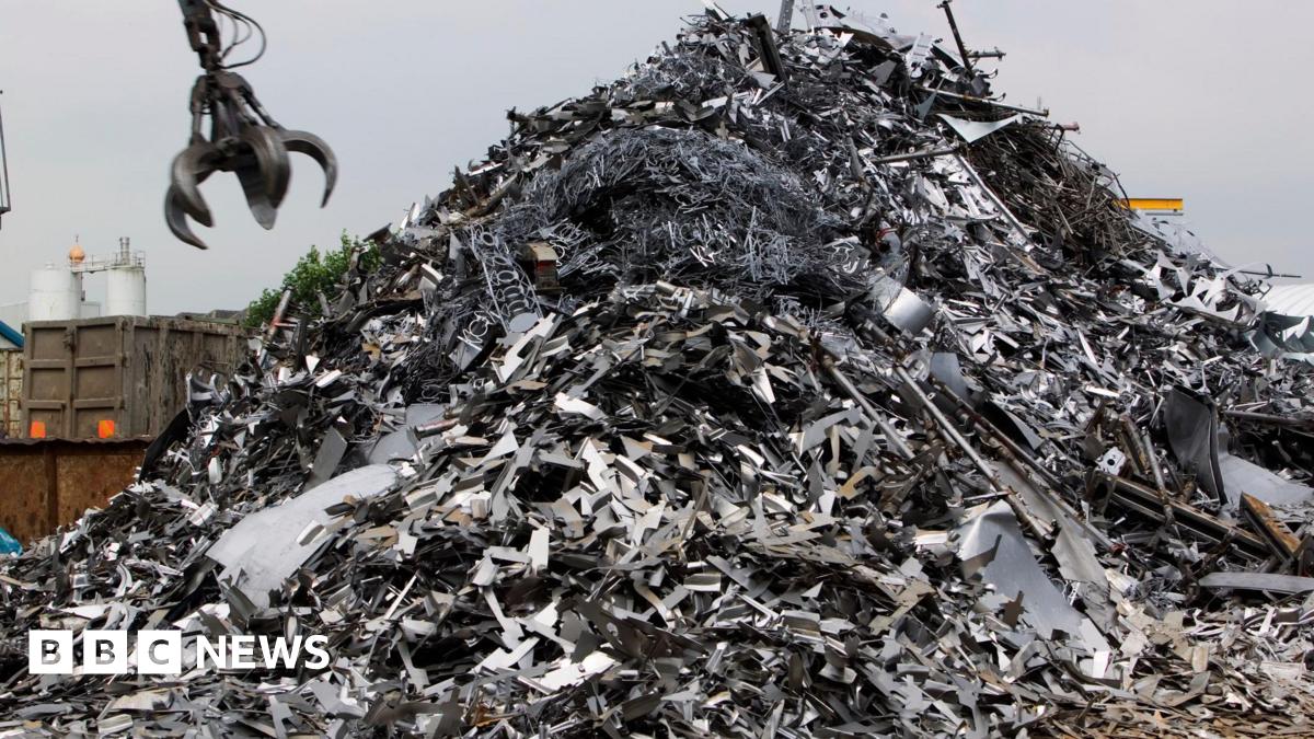 A huge pile of silver scrap metal, with a metal crane claw coming down towards it, to grab it.