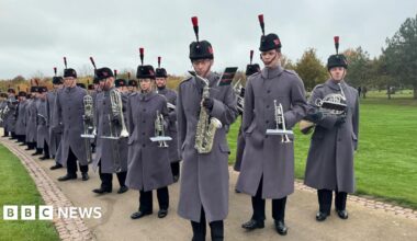 A row of soldiers in long grey coats and tall black fur hats with black feathers holding a variety of silver brass instruments on a footpath with grass on either side.