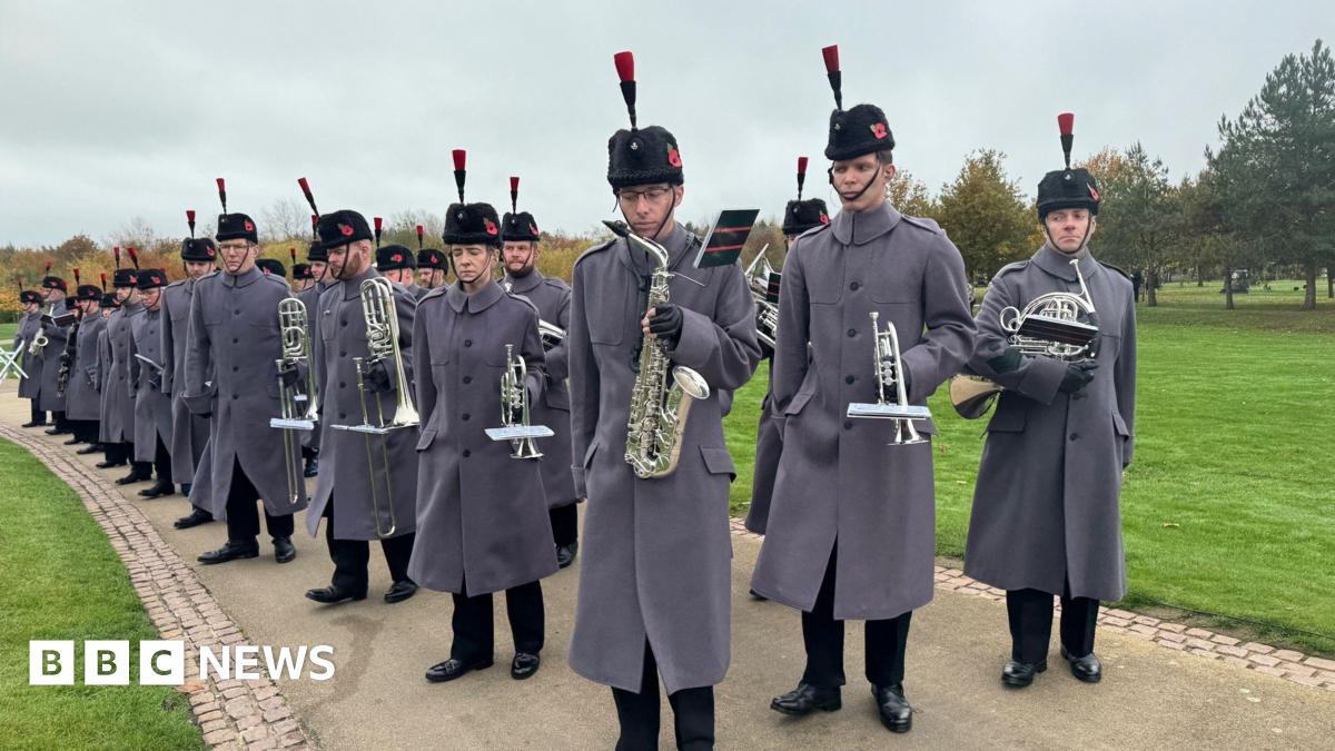 A row of soldiers in long grey coats and tall black fur hats with black feathers holding a variety of silver brass instruments on a footpath with grass on either side.