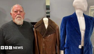 A man in a grey jumper standing next to two items of clothing. He has a grey beard and grey hair. Next to him is a brown leather jacket and a blue velvet suit which are on mannequins.