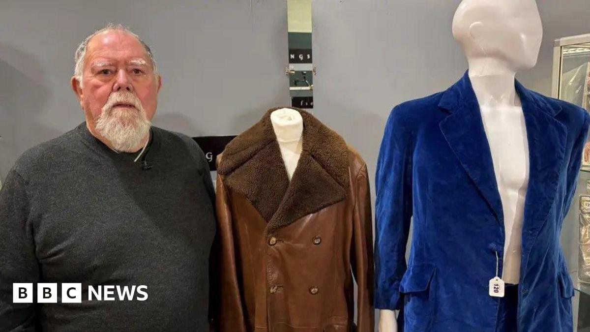 A man in a grey jumper standing next to two items of clothing. He has a grey beard and grey hair. Next to him is a brown leather jacket and a blue velvet suit which are on mannequins.