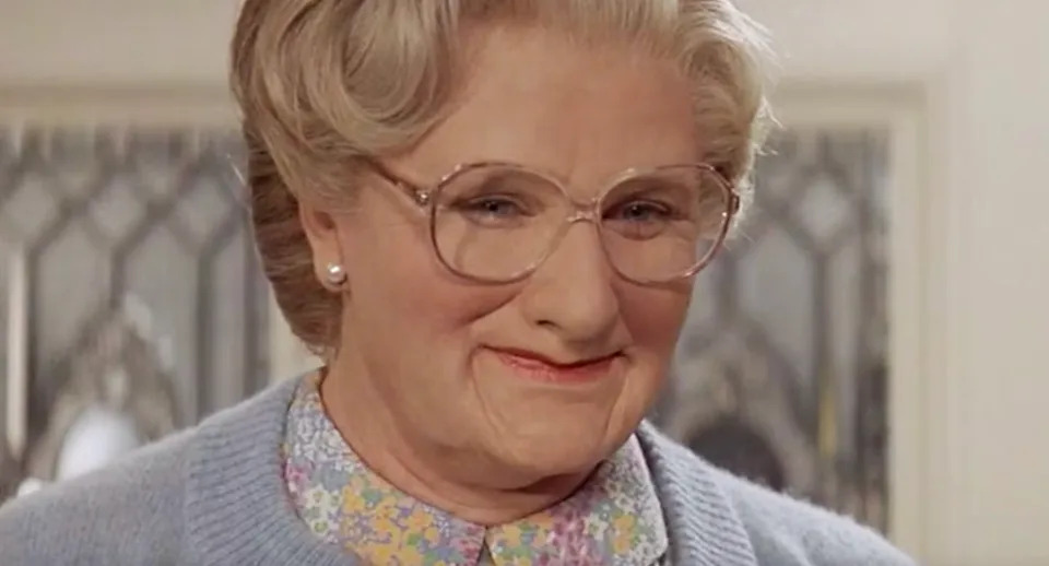 An elderly person wearing glasses, a floral shirt, and a cardigan smiles warmly