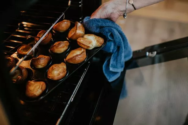 A woman uses a blue dish cloth to remove a tray full of crispy brown risen Yorkshire Puddings from an oven.