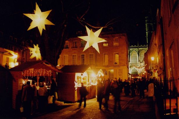 City of Bath, England hosts an annual Christmas Market, with dozens of huts selling artwork, food, drinks and other goods, all centered around citys abbey, visible in background.