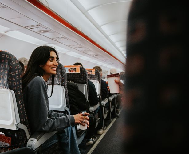 A smiling Indian woman sits in a row on a commercial aircraft. She holds a cup of coffee and looks at something off screen.