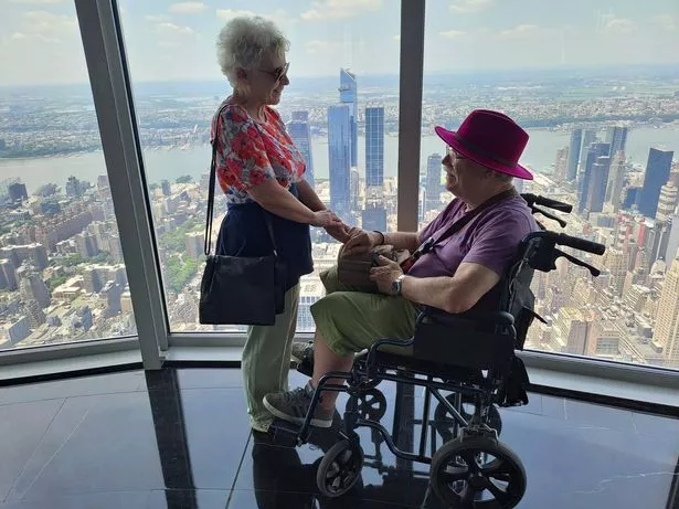 woman stood holding hands with man in wheelchair next to window with view