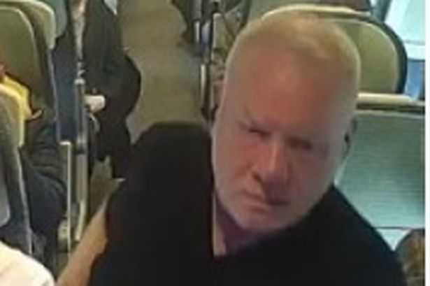 CCTV image of a man on a train who is white, clean-shaven with a shaved head, pictured wearing a black t-shirt, blue jeans and white trainers and is also carrying a phone
