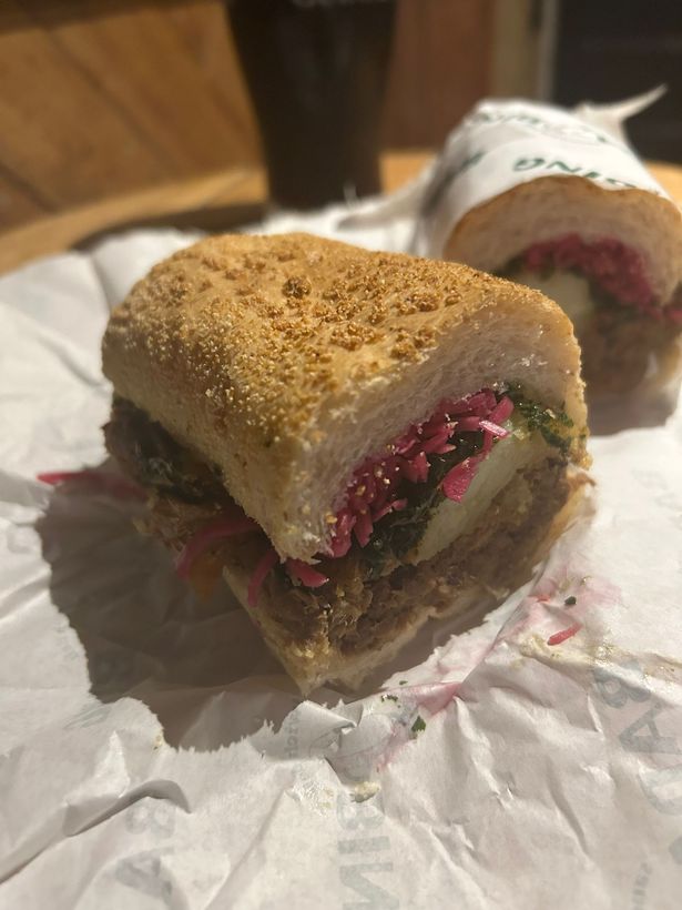 The sandwich features Guinness-soaked braised beef, crispy potatoes, pickled cabbage, crispy onions, and greens 