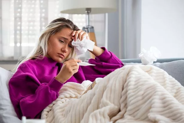Woman being sick having flu lying on sofa looking at temperature on thermometer.