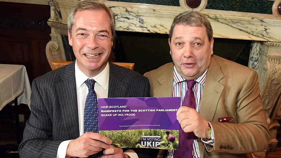 A white man with short black and grey hair wearing a pinstripe suit and a blue and white patterned tie is holding a UKIP leaflet alongside another man wearing a brown suit, stripy shirt and claret tie. They are both sat in a dining room