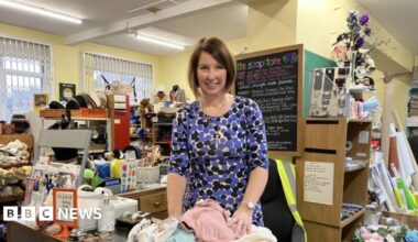 A woman in a blue and black patterned dress in a room with boxes and shelves packed with cloth and other objects