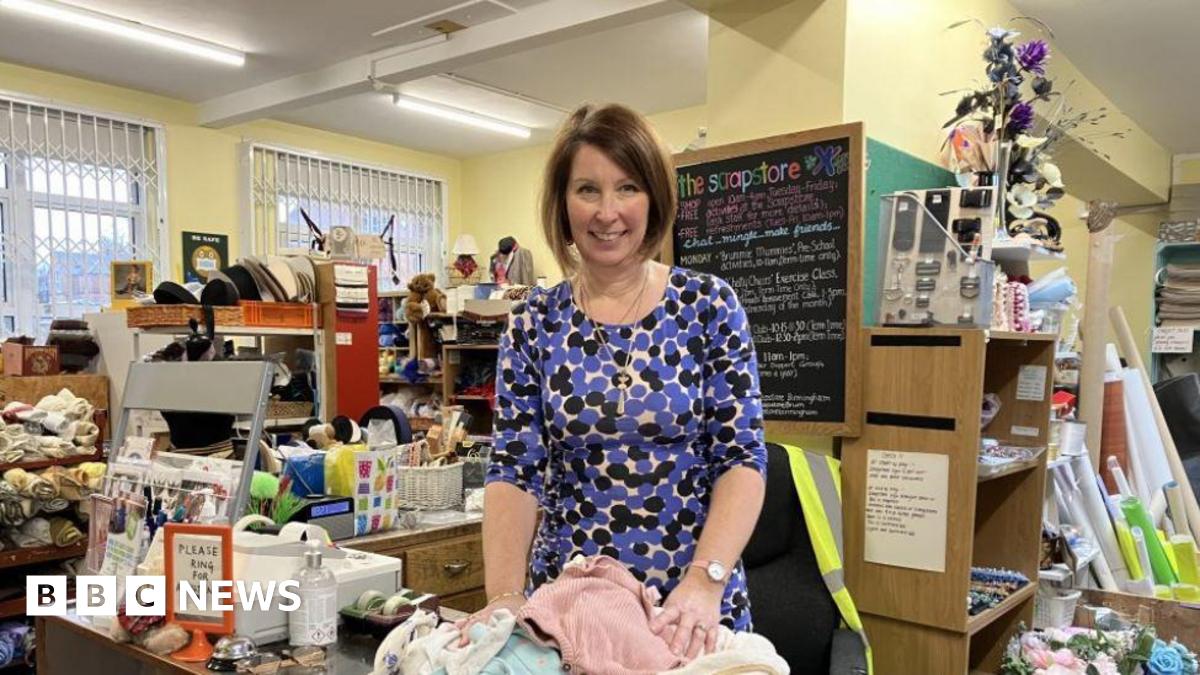 A woman in a blue and black patterned dress in a room with boxes and shelves packed with cloth and other objects
