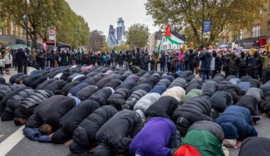 A group of Muslim men pray knelt down in the street with a crowd behind them, including someone waving a Palestine flag. Trees and the Gherkin building are visible in the skyline.