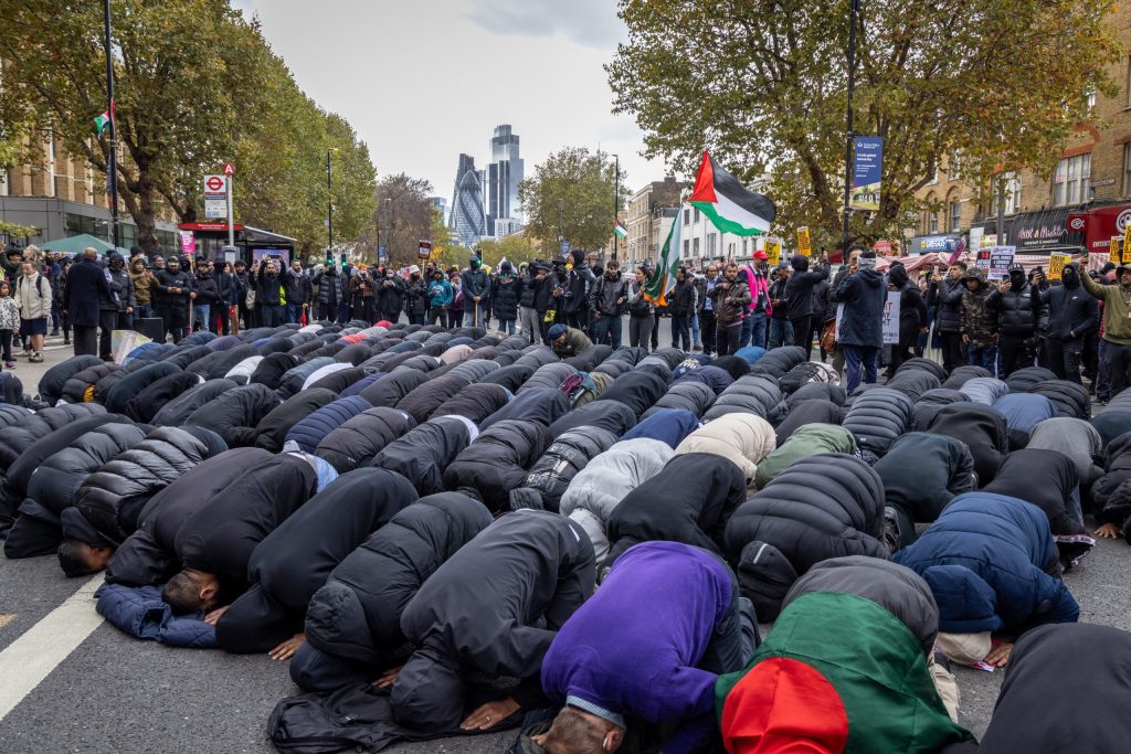 A group of Muslim men pray knelt down in the street with a crowd behind them, including someone waving a Palestine flag. Trees and the Gherkin building are visible in the skyline.