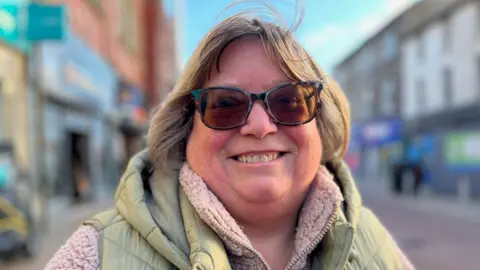Valerie Long is pictured on the high street in Rhyl. She's wearing a light green gilet with a fluffy pink fleece. She is wearing sunglasses and has a light brown bob.