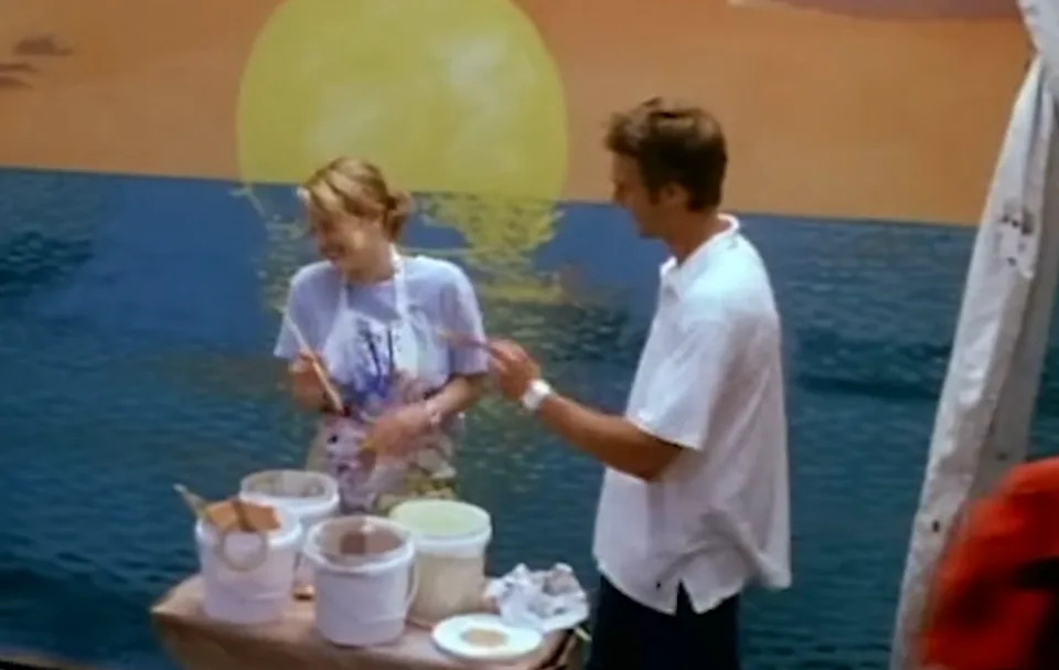 Two people engaging in a creative activity near a painted ocean mural with a sun. Several paint buckets are on a table in front of them