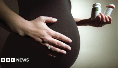 Stock image: Pregnant woman with dark top holding her stomach with her right hand and wearing a ring. In her left hand she is holding two vitamin containers.