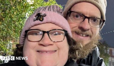 A woman with black rimmed glasses and a pink bobble hat smiles at the camera. Behind her a man with a beige bobble hat cuddles up to her. He has black rimmed glasses too and a dark beard.