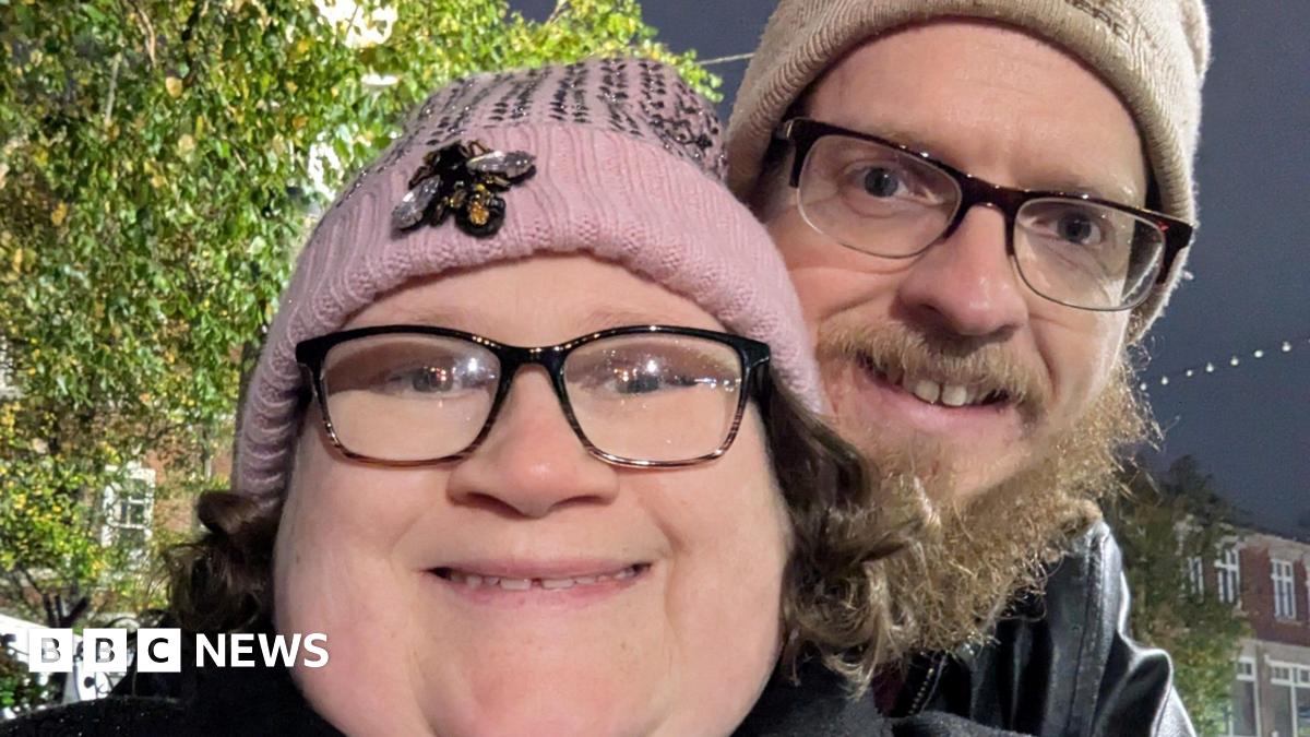 A woman with black rimmed glasses and a pink bobble hat smiles at the camera. Behind her a man with a beige bobble hat cuddles up to her. He has black rimmed glasses too and a dark beard.